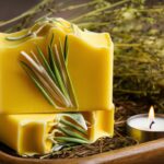 A beautifully crafted lemongrass soap, featuring a vibrant yellow hue and an intricate design of lemongrass leaves on the surface. The soap has a fresh, citrusy aroma that invigorates the senses. It sits atop a rustic wooden tray, surrounded by a bouquet of dried herbs and a small, flickering candle, creating a serene and tranquil atmosphere.