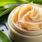 A close-up image of a rich, creamy shea butter, ready to be applied to the skin. The shea butter is a beautiful shade of ivory, with a slightly golden hue. It is displayed in a simple, elegant container, surrounded by fresh, green leaves and a few drops of water, highlighting its pure and natural origins. The overall atmosphere of the image is calming and rejuvenating.