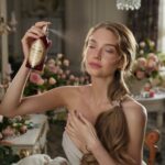 A mesmerizing image of a stunning woman spritzing pure rose water from a vintage, ornately labeled bottle onto her face. Her long, flowing hair cascades over her shoulders, and her gentle expression denotes a sense of tranquility. In the background, an elegant room filled with antique furniture, fresh flowers, and dainty chandeliers can be seen, creating an atmosphere of luxury and rejuvenation.