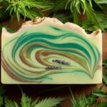 A beautifully crafted bar of hemp seed oil soap with a warm, earthy color palette. The soap is adorned with a swirling design of green and brown hues, representing the hemp seed and its natural origin. The soap has a smooth and inviting texture, and the scent of lavender is gently wafting from it. Surrounding the soap is a background of freshly harvested hemp plants, adding to the organic, eco-friendly vibe.