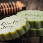 A beautifully crafted olive oil soap, featuring a soft green color with tiny flecks of gold. The soap is shaped into a rustic loaf design, with adistinctly natural and earthy scent. In the background, there is a vintage-style wooden soap dish