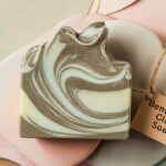 A beautifully crafted soap bar in the shape of bentonite clay, with a natural, earthy color palette. The soap features a unique swirl pattern, reminiscent of the clay's texture. The background has a calming, pastel color scheme, and there's a small tag attached to the soap with the label 'Bentonite Clay Soap'.