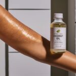 A woman's arm showcasing the benefits of coconut oil for stretch marks. Her skin glows with a healthy, radiant sheen, and the stretch marks appear reduced and smoothed out. Bottle of coconut oil displayed. Background consists of a simple, clean white backdrop.