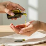 A close-up image of pure, organic avocado oil being gently poured onto a woman's hand. The hand is holding a small glass bottle of theoil, which is rich green in color. The background is minimalistic, with a light wood table and a few drops of oil on a white cloth, symbolizing the oil's effectiveness in reducing stretch marks. A soft, natural light illuminates the scene.