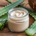 A photo of a glass jar filled with homemade face cream. The face cream is made with aloe vera and coconut oil. It has a creamy textureand is a light beige colour. The jar is placed on a wooden surface. There are fresh aloe vera leaves and a coconut in the background.